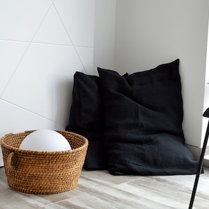 Peut inclure: Un panier en osier tressé avec une lampe blanche à l'intérieur, posé sur un sol en bois à côté de deux coussins en lin noir.
