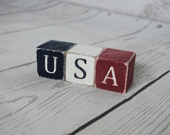 May include: Three wooden blocks arranged in a row, each featuring a distressed finish. The first block is dark blue with the white letter U, the second is white with the black letter S, and the third is red with the white letter A, spelling USA.