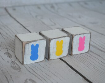 May include: Three white wooden blocks with distressed edges, each featuring a colorful bunny silhouette in blue, yellow, and pink. The blocks are arranged on a light wood-grain surface, perfect for Easter decor.