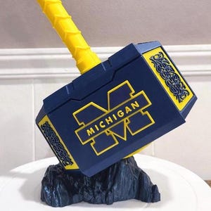 May include: A blue and yellow decorative hammer with the word "MICHIGAN" in yellow. The hammer is resting on a black rock-like base. The handle is yellow and textured.