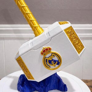 May include: A white and gold decorative hammer with the Real Madrid logo. The hammer has a gold handle and decorative gold accents. The base is blue and the hammer is on a white surface.