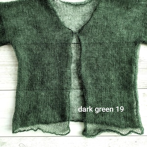 Può includere: Cardigan verde scuro con apertura frontale e maniche corte. La maglia leggera presenta una trama delicata e aperta, con un orlo smerlato. Il testo "dark green 19" è visibile. Il cardigan è presentato su una superficie di legno chiaro.