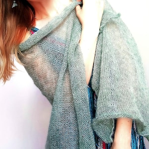 Könnte beinhalten: Ein salbeigrüner, halbtransparenter Schal in offener Strickoptik mit lockerer, luftiger Textur. Der leichte Stoff weist ein feines, zartes Maschenbild auf, das sich weich über die Schultern legt und ein durchscheinendes, geschichtetes Erscheinungsbild erzeugt.