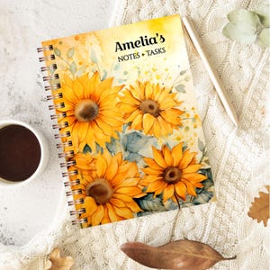 May include: A spiral-bound notebook with a sunflower watercolour design. The cover reads "Amelia's NOTES + TASKS". The notebook is open, showing the spiral binding. A pen and a cup of coffee are placed beside the notebook.