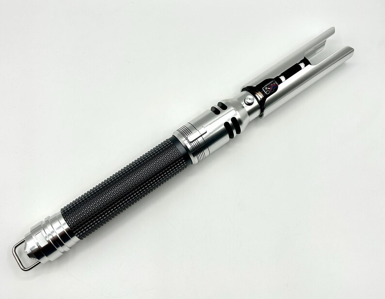 May include: A silver and black lightsaber prop with a textured black grip. The lightsaber has a silver hilt with detailed accents and a blade emitter. The prop features a loop at the base and is set against a white background.
