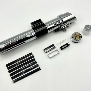 May include: A disassembled lightsaber prop with a polished chrome finish. The components include a cylindrical body, black accents, and various small parts. The design features buttons, a detailed emitter, and a gold-colored internal component. Additional black and silver pieces are also included.