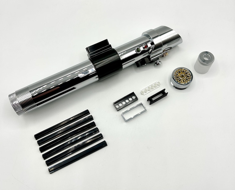 May include: A disassembled lightsaber prop with a polished chrome finish. The components include a cylindrical body, black accents, and various small parts. The design features buttons, a detailed emitter, and a gold-colored internal component. Additional black and silver pieces are also included.