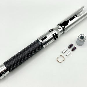 May include: A silver and black lightsaber prop with a textured black grip. The lightsaber has a silver hilt and blade emitter. Additional components, including a ring and small parts, are arranged nearby.