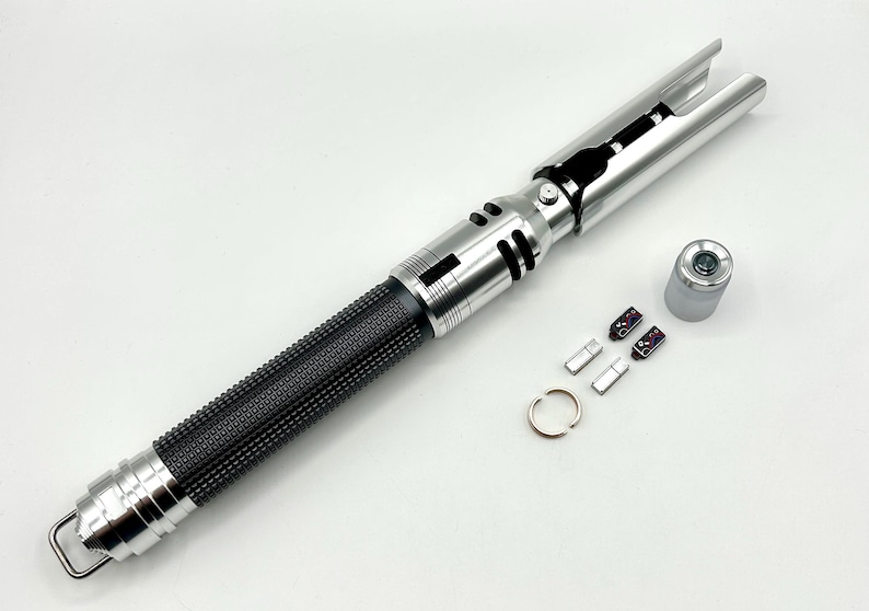 May include: A silver and black lightsaber prop with a textured black grip. The lightsaber has a silver hilt and blade emitter. Additional components, including a ring and small parts, are arranged nearby.