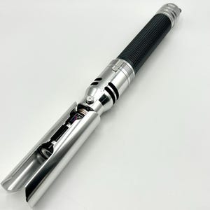May include: A silver and black lightsaber prop with a textured black grip. The hilt features intricate details and a partially exposed internal mechanism. The blade emitter is open, revealing internal components. The lightsaber is designed for cosplay or display.