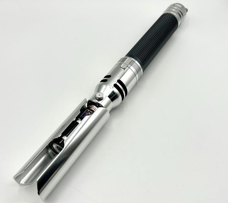 May include: A silver and black lightsaber prop with a textured black grip. The hilt features intricate details and a partially exposed internal mechanism. The blade emitter is open, revealing internal components. The lightsaber is designed for cosplay or display.