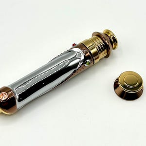 Peut inclure: Un accessoire de sabre laser métallique avec des accents argentés, dorés et cuivrés. Le manche présente un design détaillé avec des boutons et un capuchon séparé. L'esthétique générale est futuriste et détaillée, suggérant un thème de science-fiction.