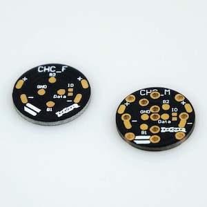 Puede incluir: Dos placas de circuito redondas negras con contactos de color dorado. La placa superior está etiquetada como "CHC_F B2" y la placa inferior está etiquetada como "CHC_M B2". Ambas placas tienen las palabras "GND", "Data", "ID" y "B1" impresas en ellas.