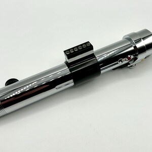 May include: A chrome-finished lightsaber prop with a black grip and control panel. The prop features a detailed design with a ribbed handle and various buttons and switches. The lightsaber is a replica of a popular science fiction weapon.