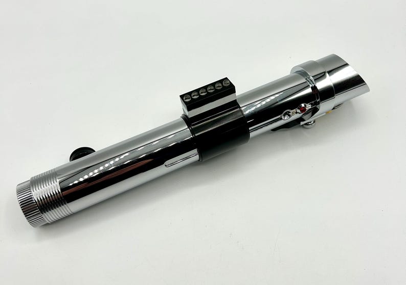 May include: A chrome-finished lightsaber prop with a black grip and control panel. The prop features a detailed design with a ribbed handle and various buttons and switches. The lightsaber is a replica of a popular science fiction weapon.
