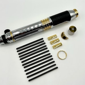 Peut inclure: Un sabre laser démonté avec un design noir, argent et or. La poignée présente une poignée noire, et les composants comprennent des accents dorés, un corps argenté et des tiges noires. Des pièces supplémentaires sont éparpillées.