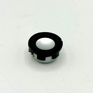 Puede incluir: Un pequeño altavoz redondo con un anillo exterior negro y un centro plateado. El altavoz tiene dos cables conectados a él.