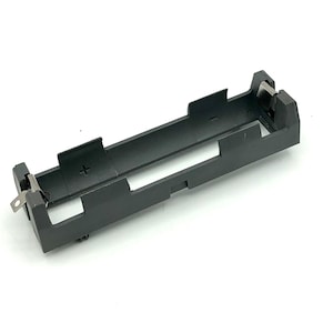 May include: A black plastic battery holder with metal contacts. The holder is rectangular with an open center and a plus sign (+) molded into the plastic. The holder is designed to securely hold a single battery.