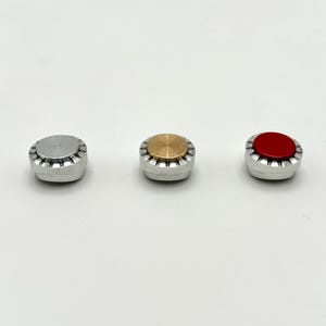 以下が含まれることがあります： 銀色のベースの3つの小さな丸いボトルストッパー。1つは銀色のトップ、1つは金色のトップ、3つ目は赤いトップです。ストッパーは白い背景に並んでいます。