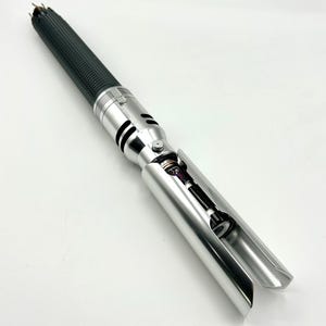 Puede incluir: Un sable de luz plateado y negro. El mango es negro texturizado y la empu&ntilde;adura es plateada. La empu&ntilde;adura tiene un bot&oacute;n y recortes negros. El emisor de la hoja est&aacute; abierto, revelando componentes internos.