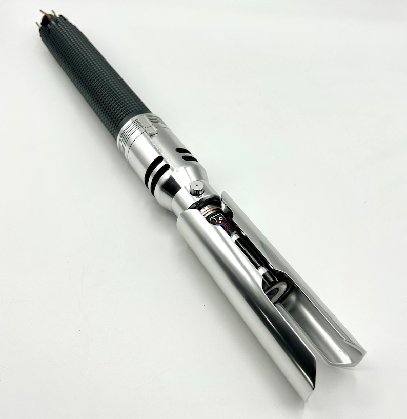 Puede incluir: Un sable de luz plateado y negro. El mango es negro texturizado y la empu&ntilde;adura es plateada. La empu&ntilde;adura tiene un bot&oacute;n y recortes negros. El emisor de la hoja est&aacute; abierto, revelando componentes internos.