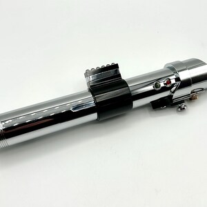 May include: A chrome-finished lightsaber prop with black accents. The prop features a cylindrical design with detailed ridges at the base and a series of buttons and lights near the top. The lightsaber is a replica of a popular science fiction weapon.