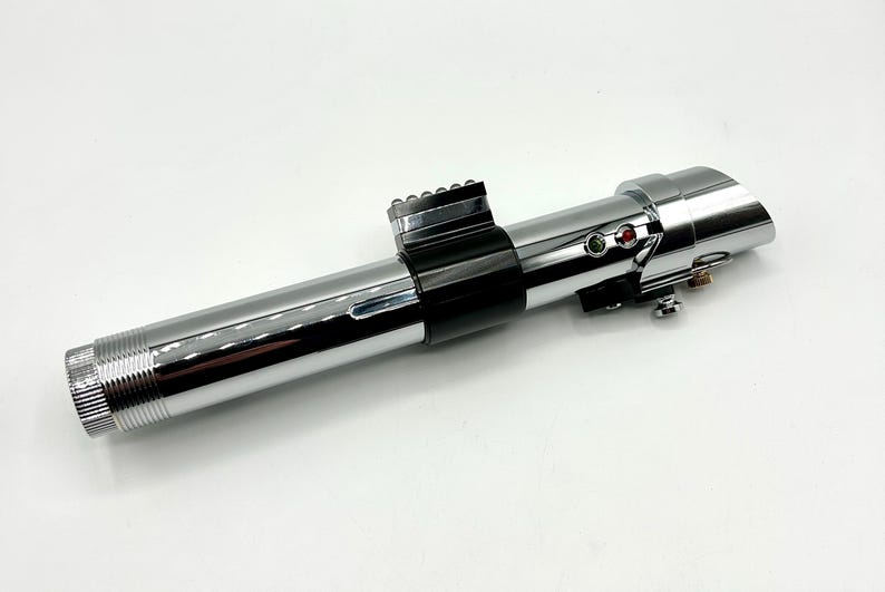 May include: A chrome-finished lightsaber prop with black accents. The prop features a cylindrical design with detailed ridges at the base and a series of buttons and lights near the top. The lightsaber is a replica of a popular science fiction weapon.