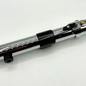 May include: A chrome-finished lightsaber prop with black accents. The prop features a cylindrical design with a ribbed handle, a black grip, and a gold-colored button. The lightsaber is a prop from a science fiction movie.