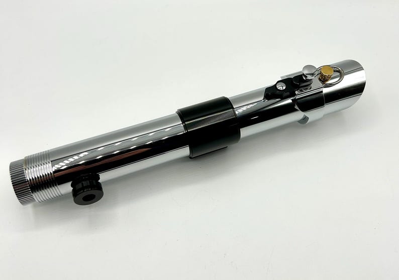 May include: A chrome-finished lightsaber prop with black accents. The prop features a cylindrical design with a ribbed handle, a black grip, and a gold-colored button. The lightsaber is a prop from a science fiction movie.