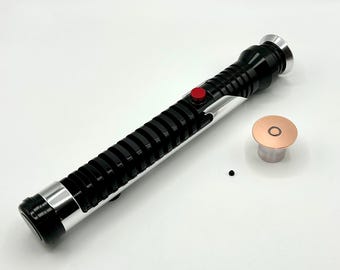 May include: A black and silver lightsaber with a red button and a separate copper-coloured piece. The lightsaber has a ribbed design and silver accents. The copper piece has a circular design in the centre.