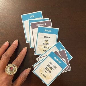 May include: A collection of blue and purple cards with white text, including the words "SITA", "HANUMAN", and "RAVAN". The card "RANGOLI" is also visible. A gold ring with a red stone is on a hand.