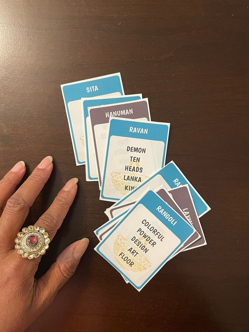 May include: A collection of blue and purple cards with white text, including the words "SITA", "HANUMAN", and "RAVAN". The card "RANGOLI" is also visible. A gold ring with a red stone is on a hand.