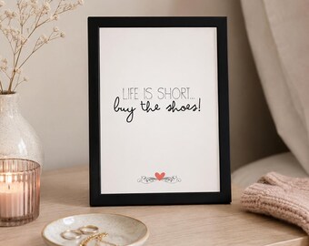 May include: A black rectangular picture frame containing a white print with the black text "LIFE IS SHORT... buy the shoes!" and a small red heart graphic centered at the bottom. The frame is positioned upright on a light wooden surface.