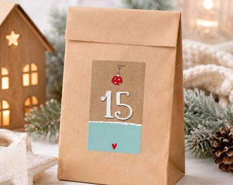 May include: A brown paper bag featuring a rectangular label with the number 15 in white, a small red Christmas ornament illustration, and a light blue bottom section with a tiny red heart, centered on the front of the bag.
