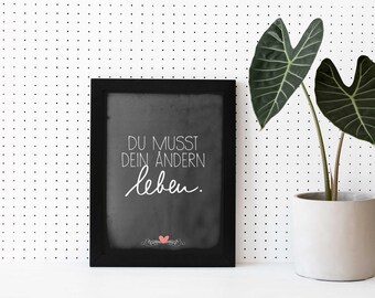 May include: Black framed chalkboard with white text that reads "Du musst dein andern leben."  The chalkboard is on a white background with a black and white polka dot pattern. A green plant with large leaves is in a white pot to the right of the chalkboard.