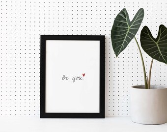 May include: A black framed print with the text "Be you." in cursive with a small red heart above the "u". The print is sitting on a white surface with a potted plant in the background.