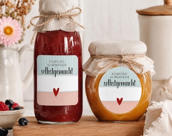 May include: A tall glass bottle filled with red jam and a round glass jar filled with orange jam, both topped with beige fabric covers tied with twine. Each container features a label with the text KLEINER GRUSS AUS MEINER KÜCHE selbstgemacht.