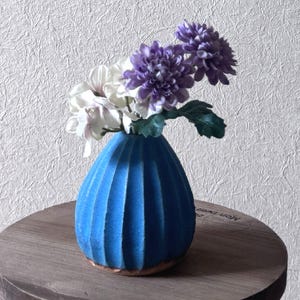以下が含まれることがあります： 縦溝のある青い花瓶には、白と紫の花のブーケが入っています。花瓶は小さくて丸い木製のテーブルの上に置かれています。背景はテクスチャ加工された白い壁です。