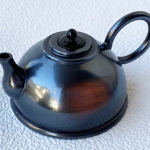 Puede incluir: Una tetera pequeña de metal negro con cuerpo redondeado y un pomo negro en la tapa. La tetera tiene un acabado metálico brillante y un aspecto ligeramente desgastado.