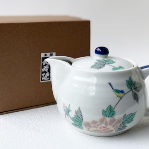 May include: A white ceramic teapot with a blue and green floral design and a small bird. The teapot has a lid and a handle. The teapot is sitting on a brown box with Japanese text.