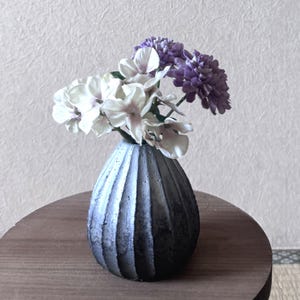 以下が含まれることがあります： 縦の溝が入った小さな濃い灰色の花瓶に、白と紫の花のブーケが入っています。花瓶は丸い木製のテーブルの上にあります。背景は、テクスチャ加工されたオフホワイトの壁です。