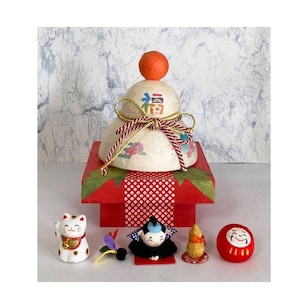 May include: A white paper mache Japanese New Year's mochi cake with a red and gold ribbon and a small orange ball on top. The mochi is on a red and white patterned box with a green base. There are five small figurines in front of the mochi, including a white cat, a black cat, a gold cat, a red daruma doll, and a gold figurine.
