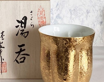Könnte beinhalten: Eine goldene Teetasse aus Keramik mit einer strukturierten Oberfläche. Die Tasse steht auf einer weißen Oberfläche neben einer Holzkiste mit japanischen Schriftzeichen.
