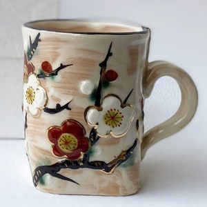 May include: A ceramic mug with a cream-colored background and a floral design. The flowers are white, red, and yellow with gold accents. The mug has a black rim and a handle.