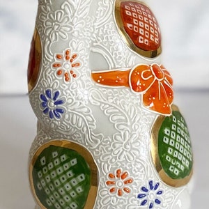 May include: White ceramic figurine of a Japanese Maneki-Neko, or lucky cat, with gold accents and colorful floral patterns. The cat is sitting with its right paw raised, a traditional gesture for good luck and fortune.