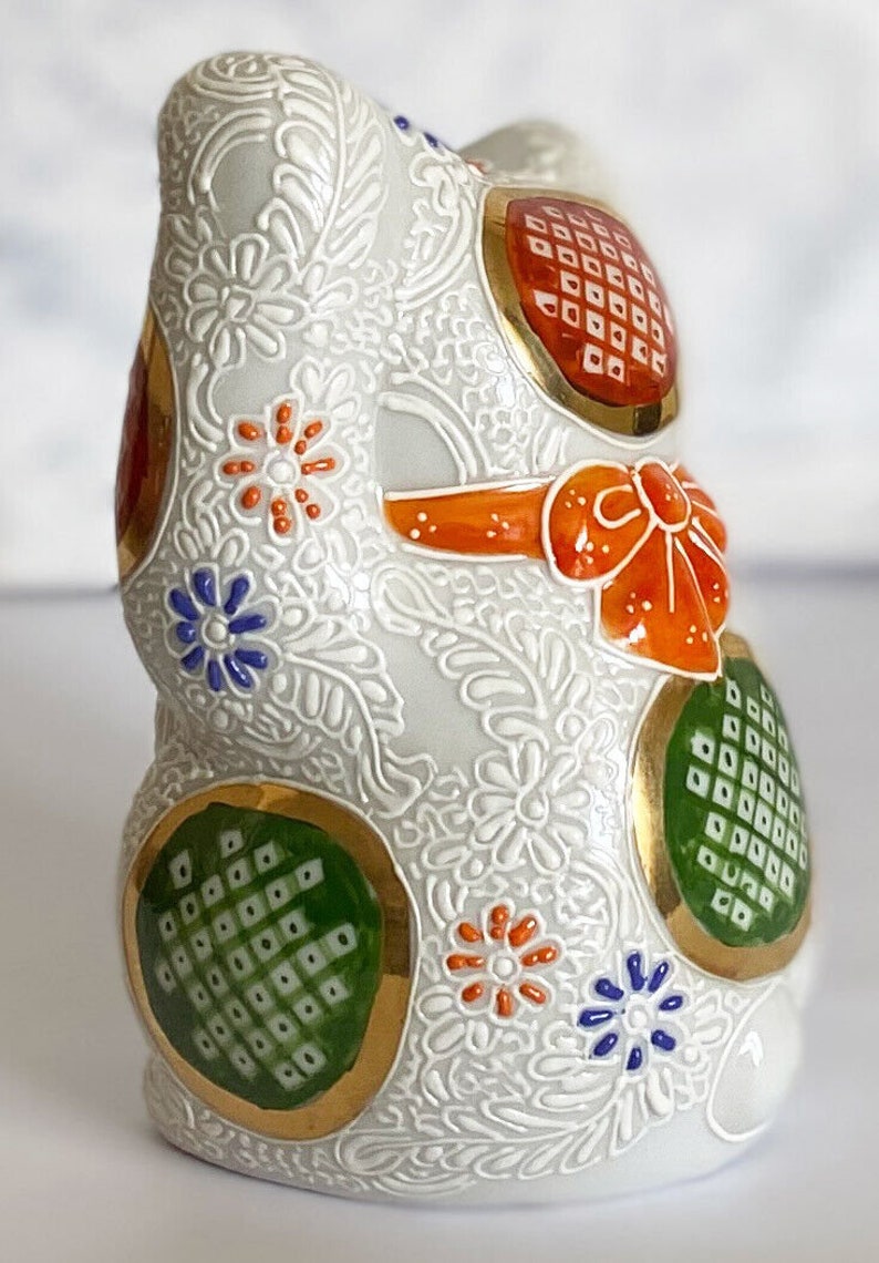 May include: White ceramic figurine of a Japanese Maneki-Neko, or lucky cat, with gold accents and colorful floral patterns. The cat is sitting with its right paw raised, a traditional gesture for good luck and fortune.