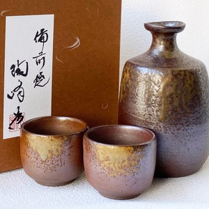 Op de afbeelding: Een set van drie bruine en gouden keramische sake-waren, waaronder een fles en twee bekers. De fles heeft een getextureerd oppervlak en de bekers hebben een gespikkelde glazuur. De set wordt geleverd in een bruine doos met Japanse kalligrafie.