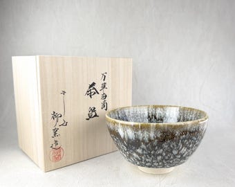 May include: A ceramic tea bowl with a dark brown and white mottled glaze, featuring a lighter rim. Beside it sits a light-colored wooden box with black Japanese calligraphy characters and a red circular stamp on the front surface.