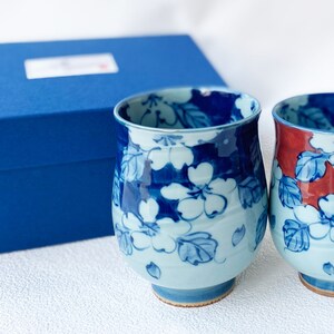 May include: Two blue and white ceramic teacups with floral designs. One cup has a red accent. The cups are in a blue gift box.