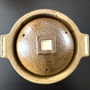 May include: A brown ceramic pot with a lid, featuring a textured surface and a small square opening in the center of the lid.